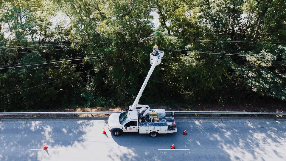 Aerial view of a utility worker on a bucket truck fixing power lines by a road.