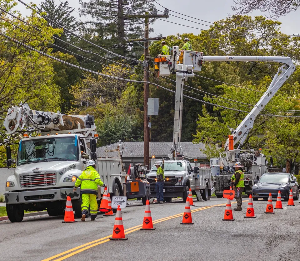 Utility workers in reflective gear repair power lines using bucket trucks and cones on a suburban street.