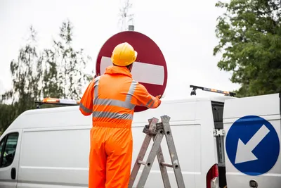 A road worker in orange uniform adjusting a traffic sign on a ladder by a van.